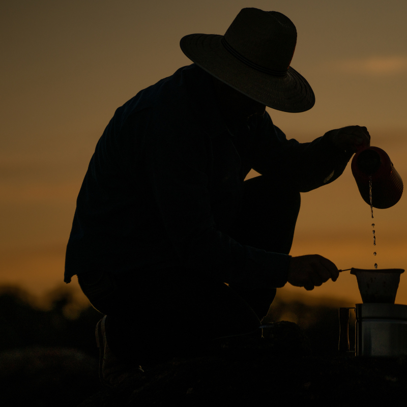 Silhouette of a person in a wide-brim hat kneeling at sunset, pouring water from a kettle into a pour-over coffee dripper, warm amber sky in the background.