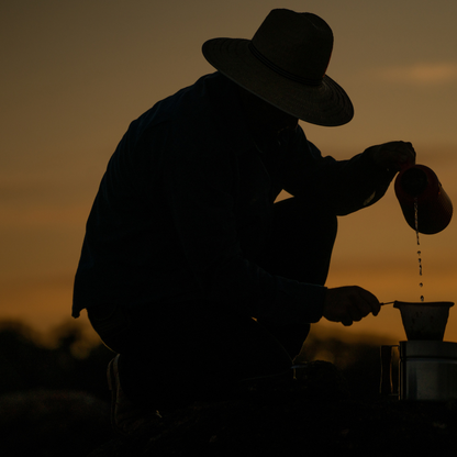 Silhouette of a person in a wide-brim hat kneeling at sunset, pouring water from a kettle into a pour-over coffee dripper, warm amber sky in the background.
